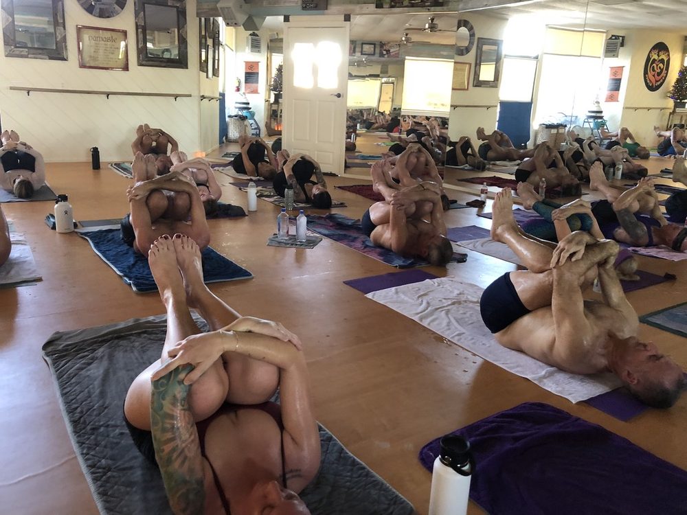 Bikram Yoga El Paseo class practicing knees-to-chest pose on yoga mats in hot yoga room.